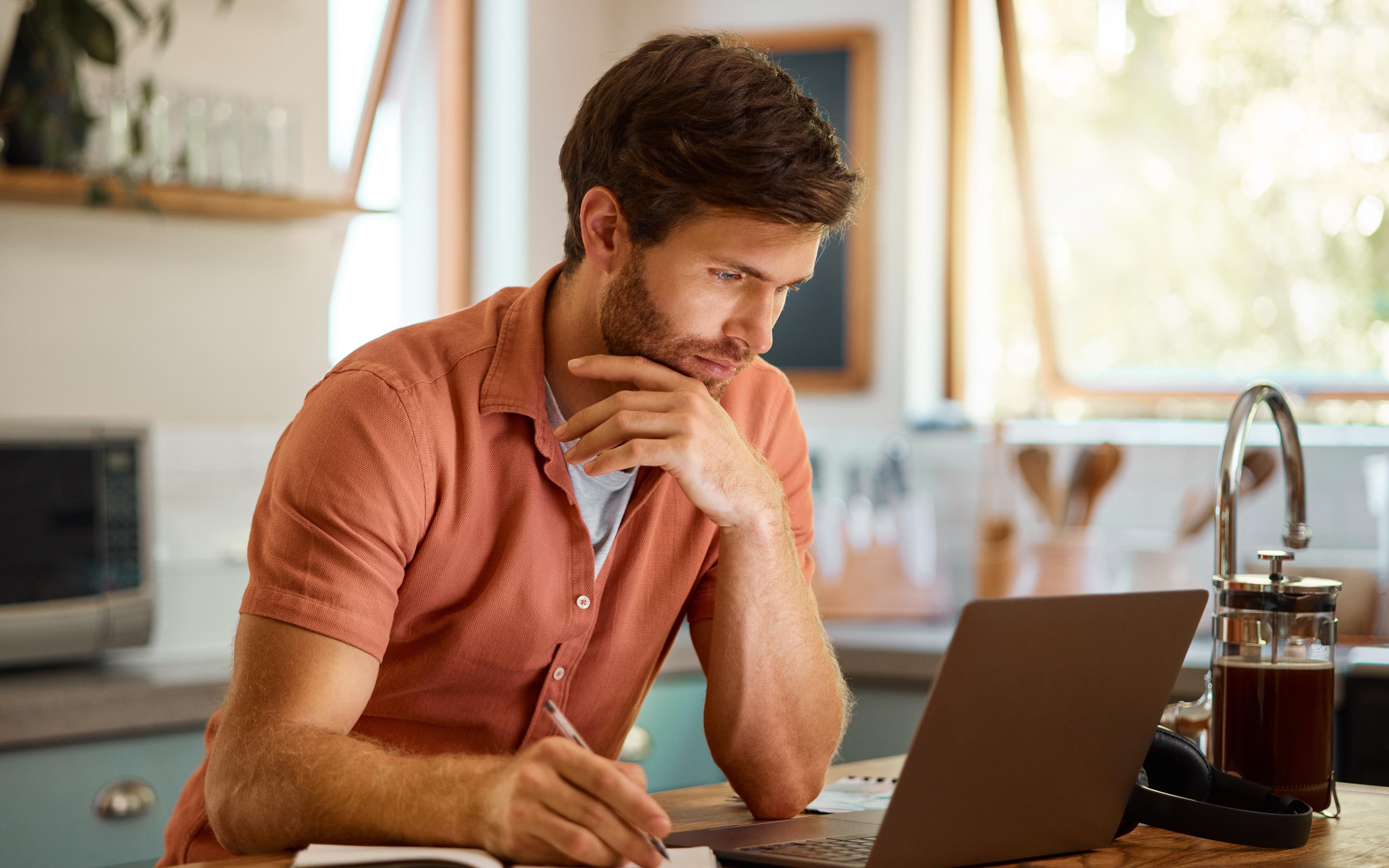 Homeowner sitting at a kitchen table using a laptop to research window repair options for his home near a sunlit window